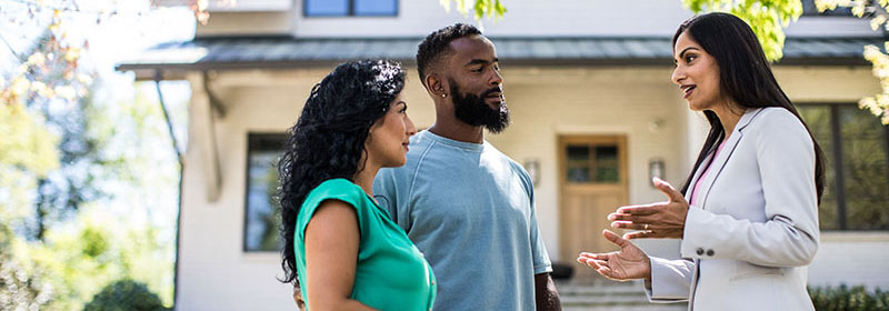 A young couple discusses low down payment options with a realtor outside a new home 