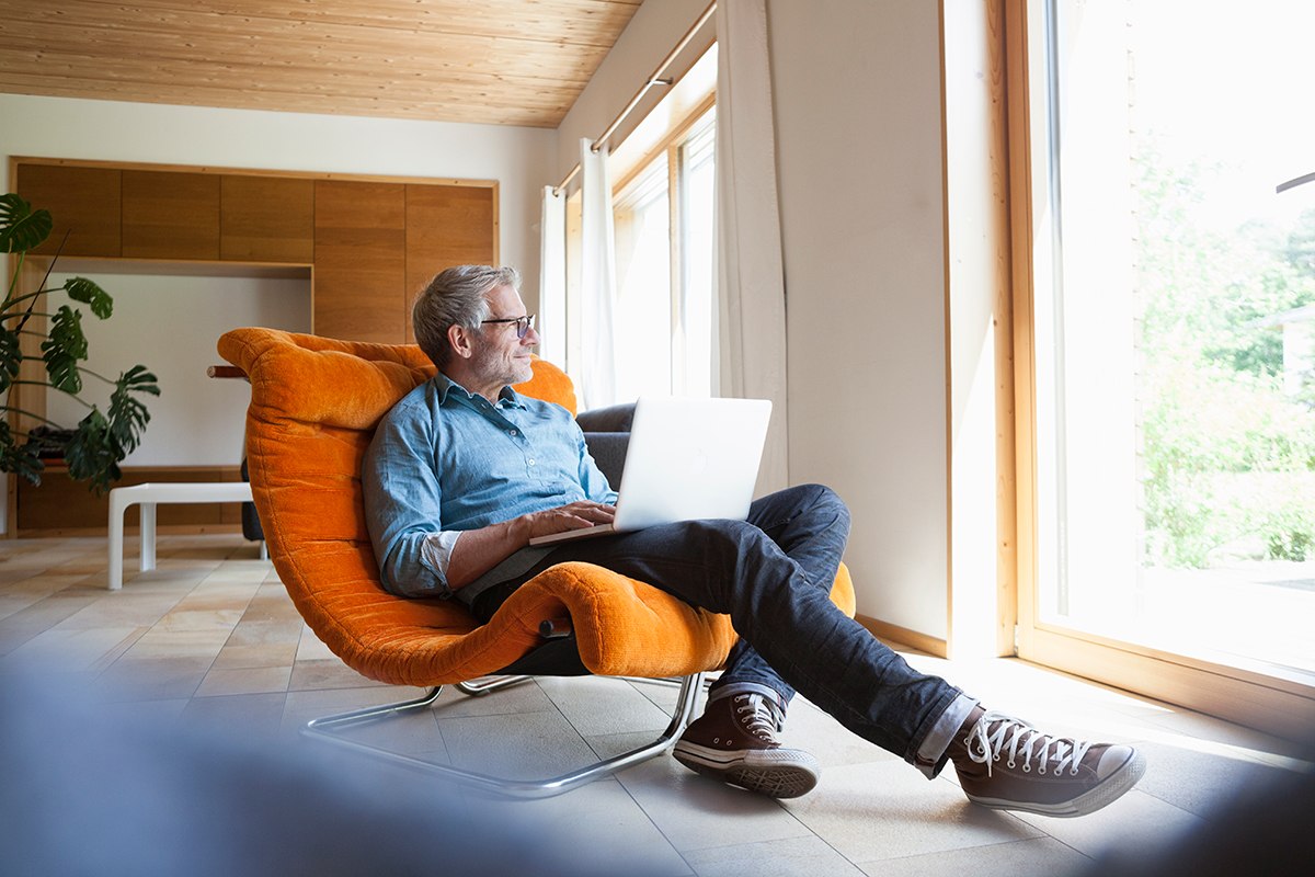 A man sitting in a orange chair, with a laptop on his lap looking outside