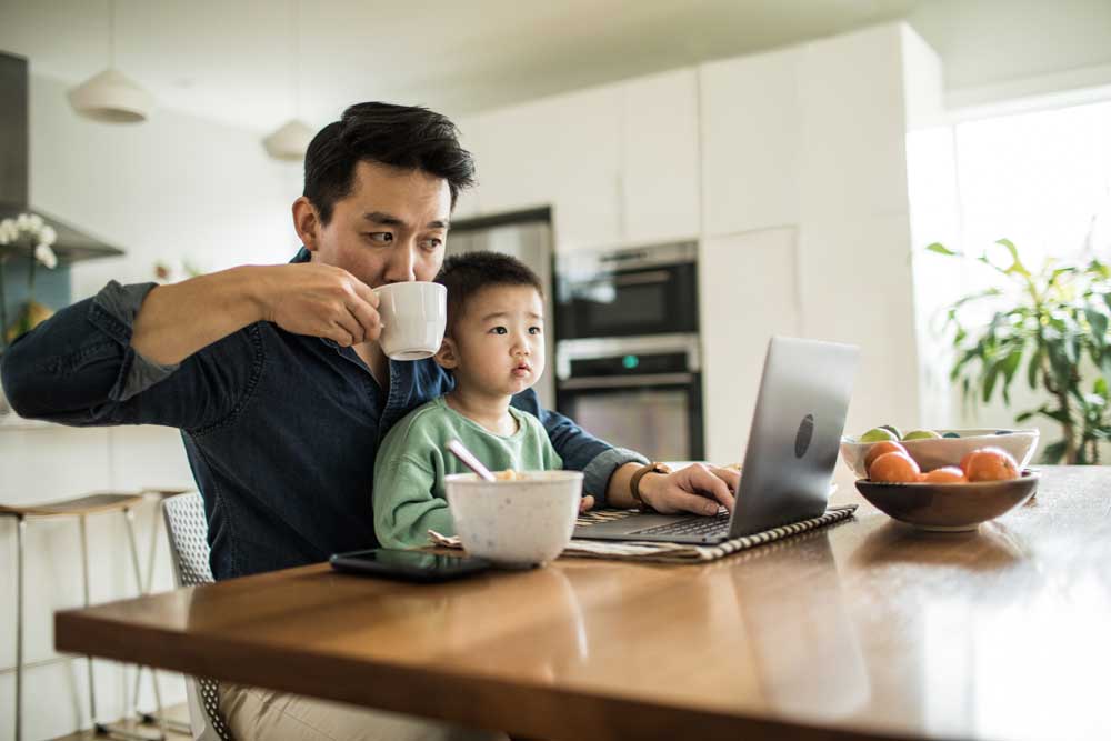 a man looks at his laptop with his son sitting in his lap