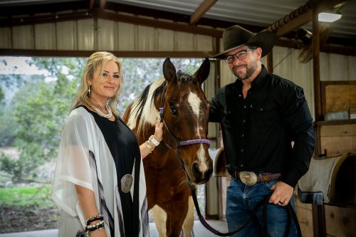 A stylish couple posing with a horse in a barn setting, showcasing their western attire and accessories.