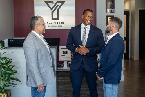 Three professionals engaged in conversation in an office setting, with a logo for Yantis prominently displayed in the background.