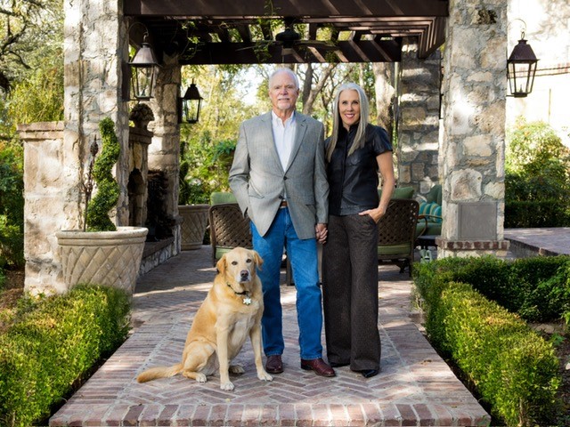A couple standing on a stone patio walkway under a pergola, with their golden retriever sitting in front.