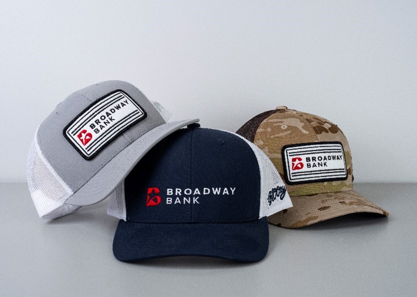 Three stylish baseball caps featuring the Broadway Bank logo, showcasing different designs including a gray cap, a navy cap, and a camo cap.