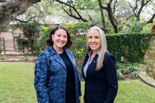 Two women smiling outdoors in a garden with trees and greenery in the background.