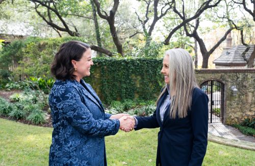 Two women shaking hands outdoors in a garden near a stone gate.