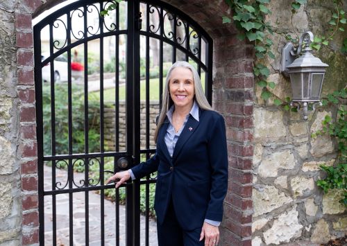Woman in a navy suit standing by a wrought-iron gate in a stone archway with ivy and a wall lantern.