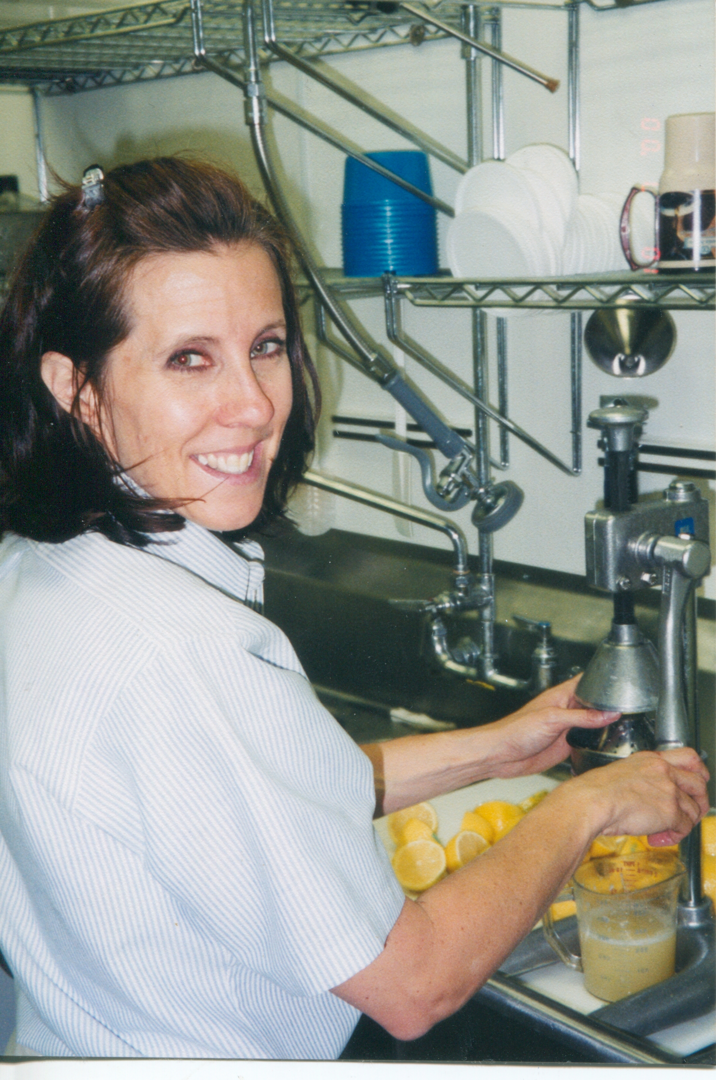 Smiling woman using a manual citrus juicer at a commercial kitchen sink, with cut lemons nearby.