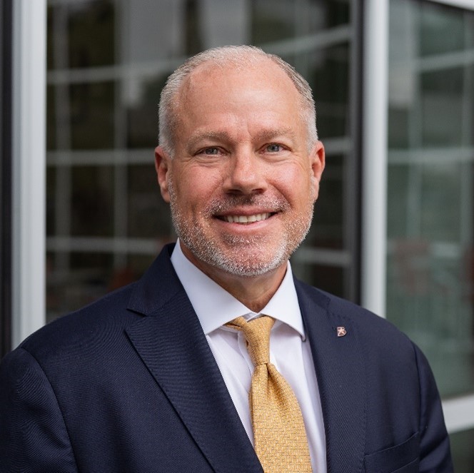 Professional portrait of a man in a suit with a light tie, smiling confidently against a modern background.