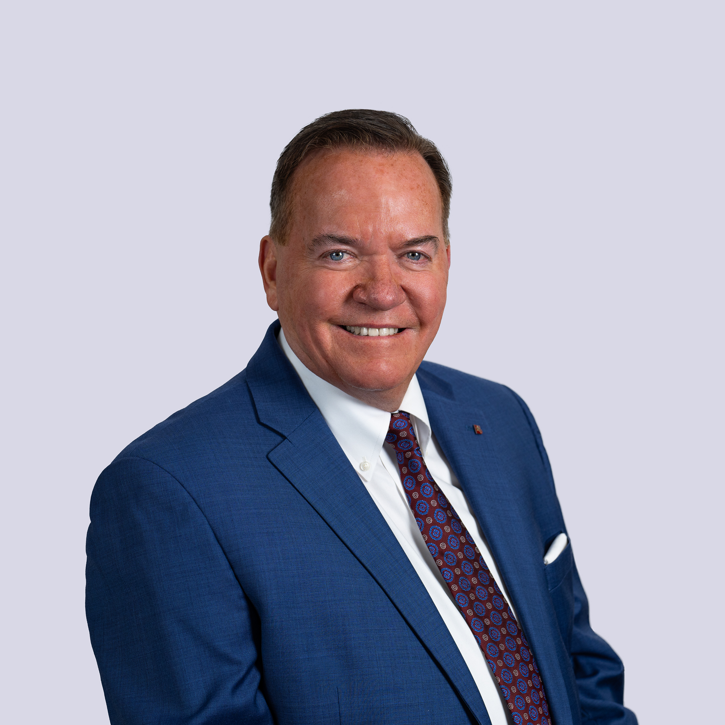Professional headshot of a man in a blue suit with a tie, smiling against a neutral background.