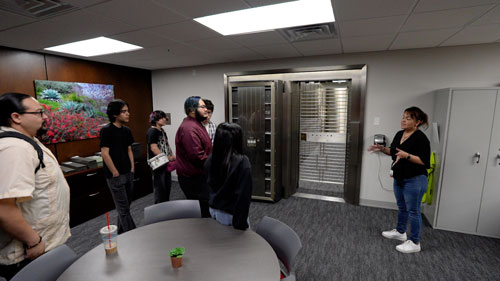 A group of seven people attentively listens to a speaker in a modern office setting, near a secure vault entrance.