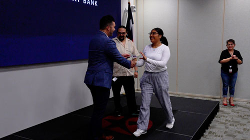 A woman is receiving an award from a man during a recognition event at a bank, with other attendees in the background.
