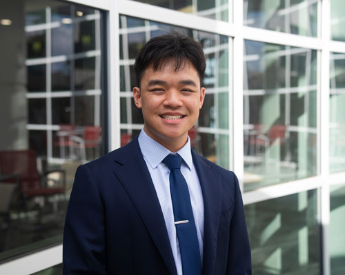 Smiling young man in a suit stands in front of a modern glass building.