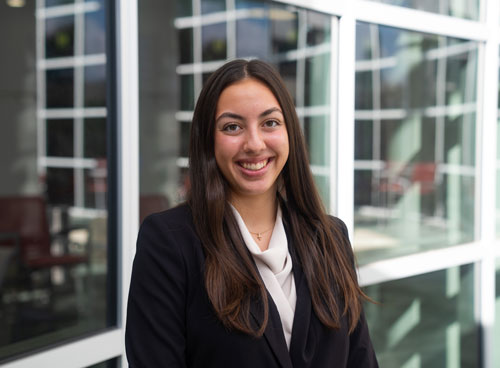 A professional portrait of a young woman wearing a black suit and smiling in an office setting with large windows in the background.
