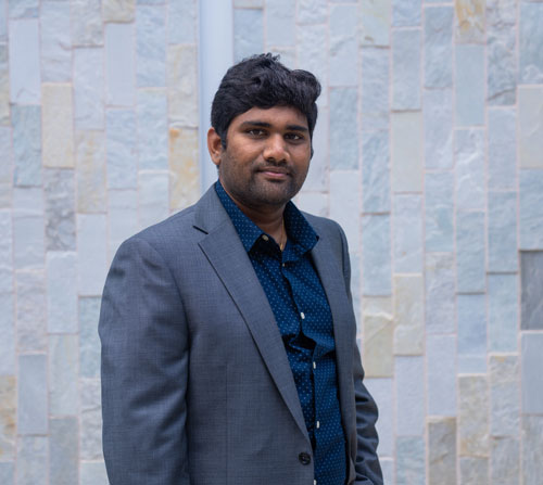 A man dressed in a formal suit stands confidently against a textured stone wall.