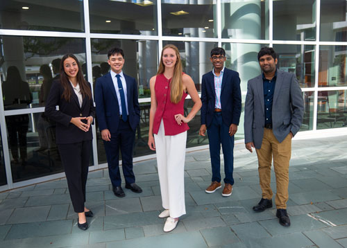 Group of five professionals posing outdoors in business attire, smiling and looking towards the camera.