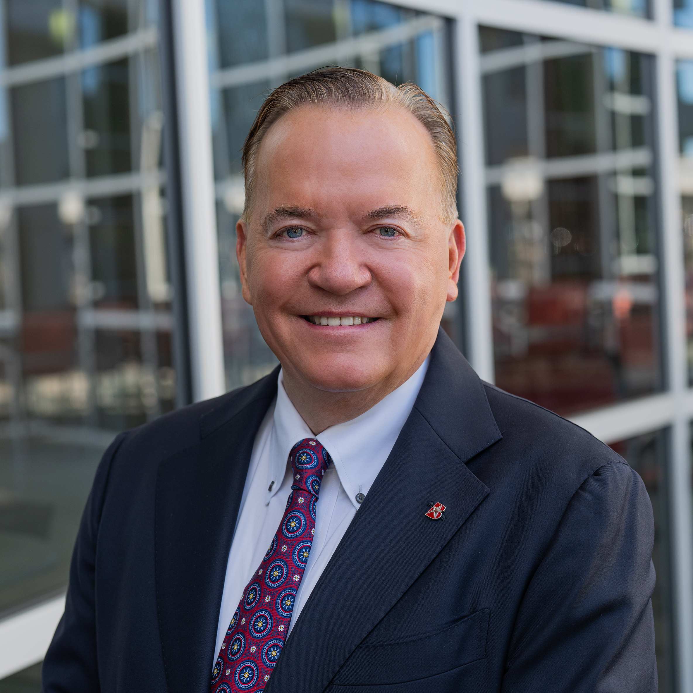 Smiling man in a navy suit and patterned tie standing in front of a modern glass building.