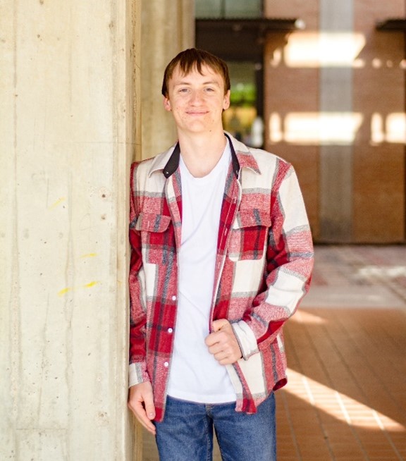 A young man in a stylish red and white plaid shirt leans against a concrete pillar, exuding a relaxed and confident vibe.